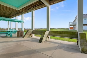 An outdoor covered area with benches and picnic tables, a trash can, and a view of the beach and ocean. - Gabriels Landing 5 Bedroom Home (Port Bolivar)