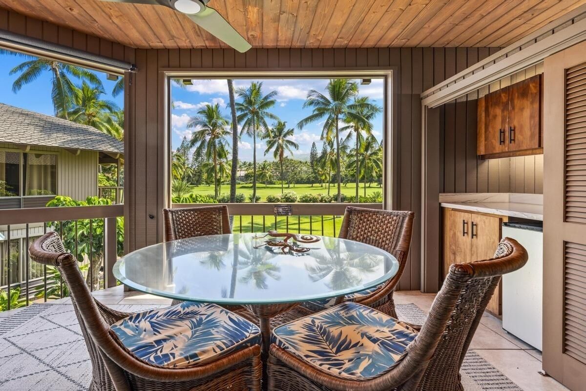 A round glass table with wicker chairs, a wooden ceiling, and a view of palm trees.