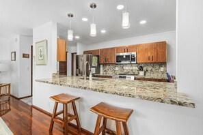 A modern kitchen with wooden cabinets, granite countertops, stainless steel appliances, and pendant lighting. - Big Island Kanaloa At Kona 803 1 Bedroom Condo (Kailua-Kona)