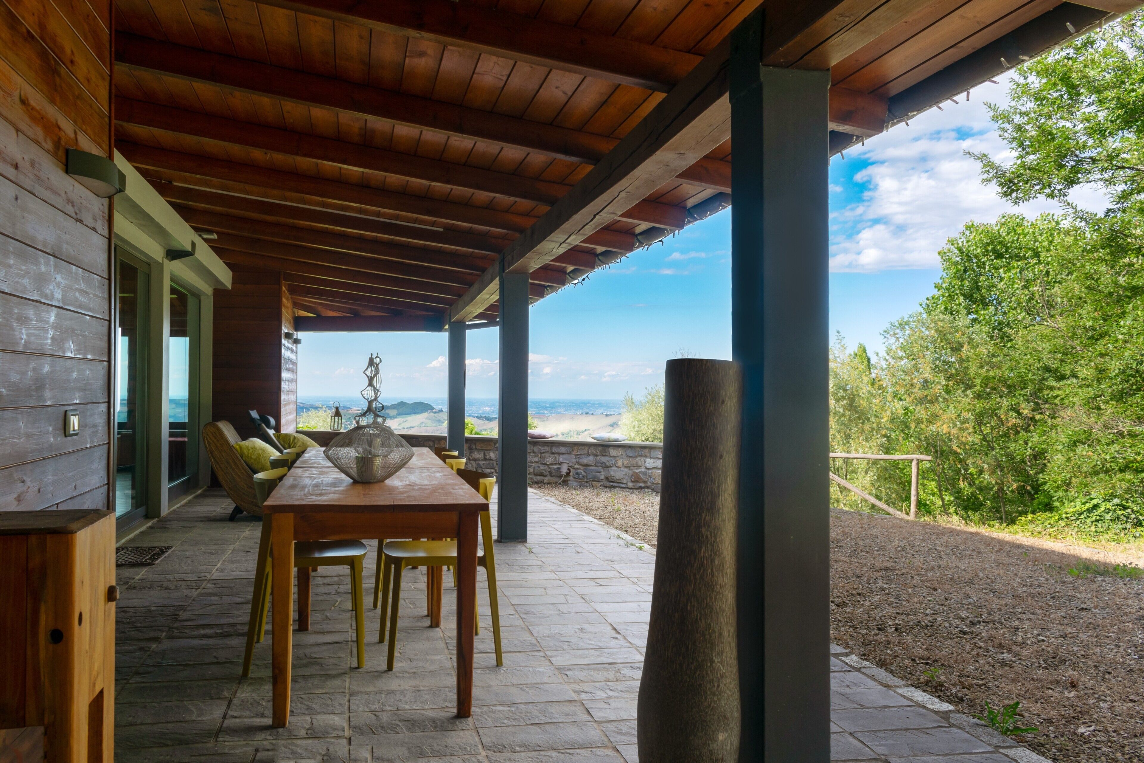 An outdoor patio with a wooden table and chairs, a stone floor, and a view of the landscape.