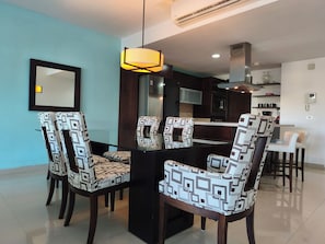 A modern dining area with a central table surrounded by patterned chairs, a hanging light fixture, and a kitchen in the background. - Ocean View Condo with Private Splash Pool Steps from the Beach at Mareazul (Playa Del Carmen)