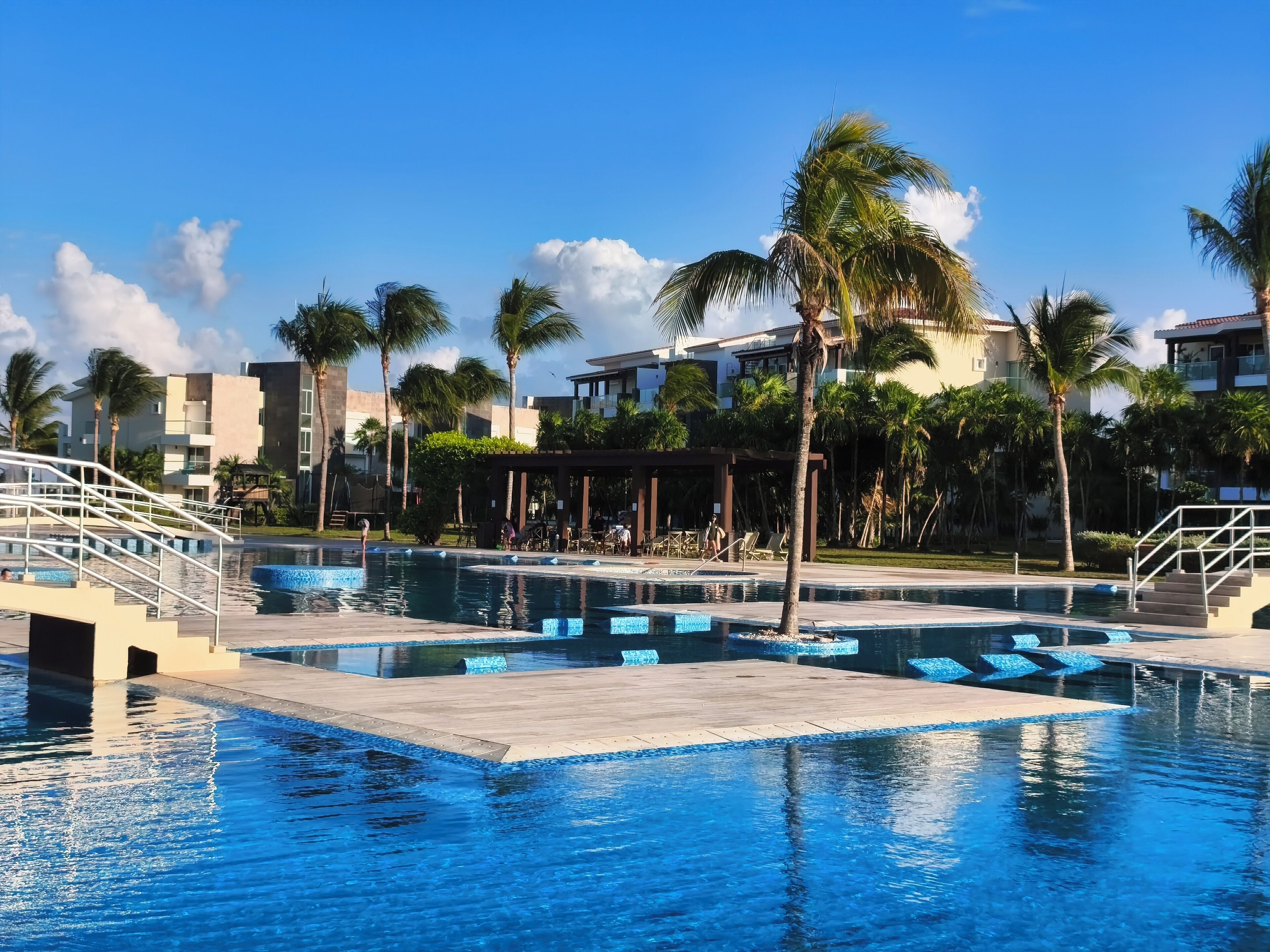 A resort pool area with multiple swimming pools, palm trees, and a covered seating area.