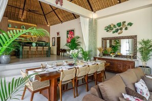 A dining area with a long wooden table set for six, surrounded by wicker chairs, with a large mirror and decorative leaves on the wall. - 3-bedroom Villa in beautiful Tabanan with rice field and river view (tabanan)