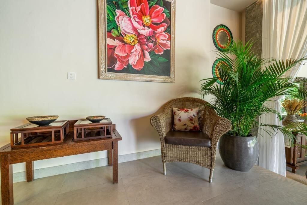 A living room with a wicker sofa, a coffee table with bowls, a large floral painting, and a potted plant.
