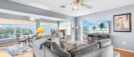A spacious living room with a gray sectional sofa, a dining area with a yellow lamp, and a large window offering a view of the outdoors.