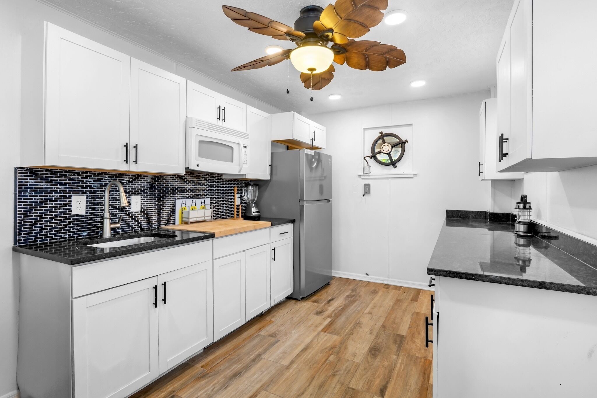 Una cocina moderna con armarios blancos, encimera negra, suelo de madera y un ventilador de techo.