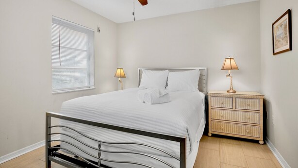 A bedroom with a bed, bedside lamps, a ceiling fan, a window with blinds, and a bedside table with a lamp. - Island Bay House B in Anna Maria Island (Holmes Beach)