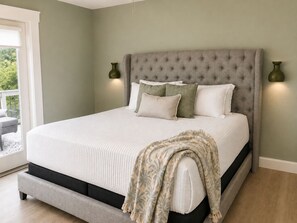 A bedroom with a large bed, a grey upholstered headboard, two wall-mounted lamps, and a window with a view of greenery. - Private heated pool & hot tub near AMI and IMG (Bradenton)
