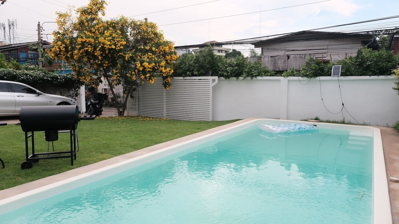 A rectangular swimming pool with a white border, a grill, and a tree with yellow flowers.