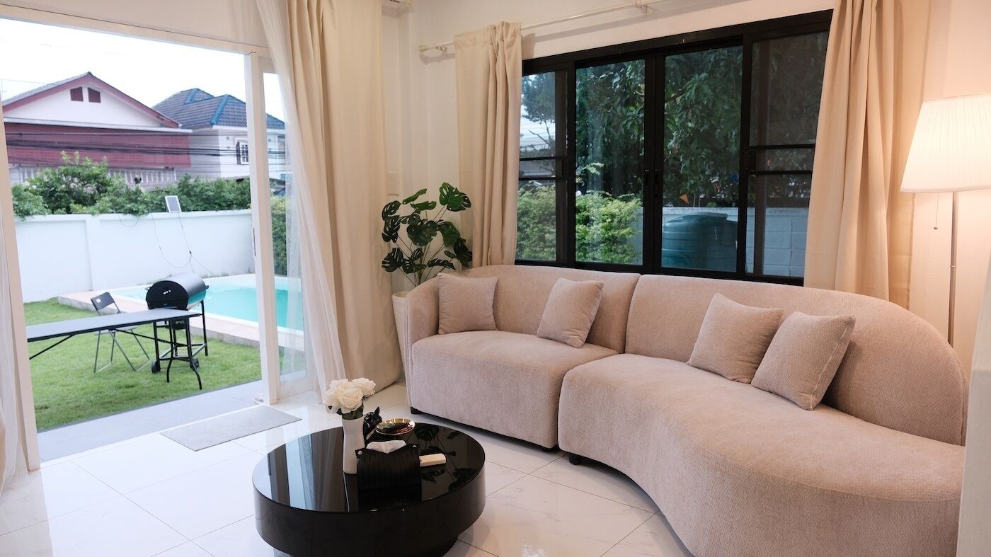 A living room with a curved sofa, a glass coffee table, and a view of a backyard with a barbecue and pool.