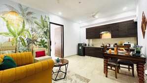 Una sala de estar con un sofá amarillo, una mesa de comedor con sillas, una zona de cocina con armarios oscuros y un mural con plantas tropicales en la pared.