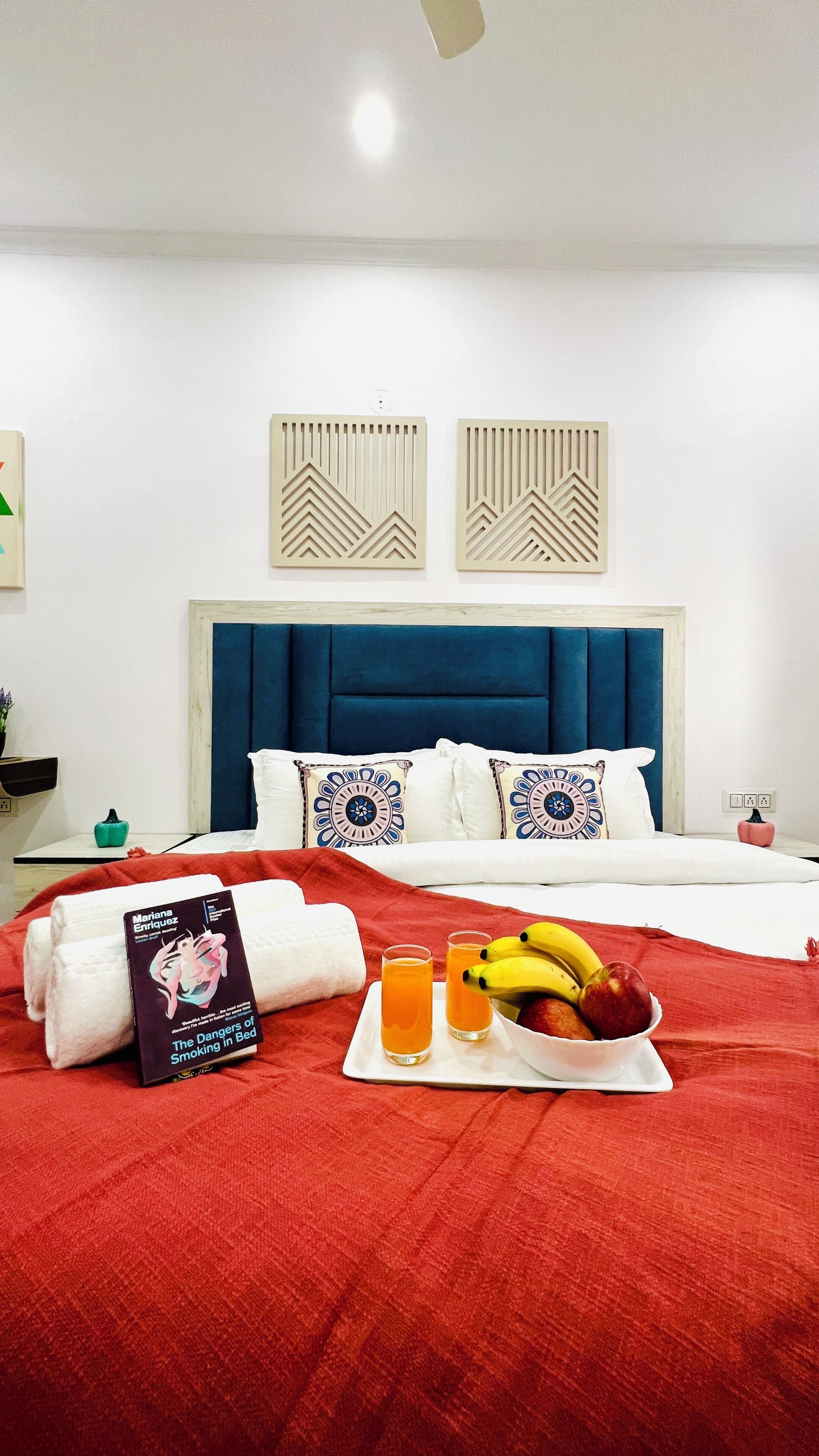 A neatly made bed with a red blanket, a book titled 'The Dangers of Smoking in Bed,' two glasses of orange juice, and a bowl of fruit on the bed.