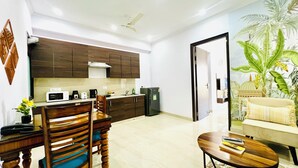 Una cocina moderna con armarios de madera oscura, un microondas y un refrigerador. Hay una mesa de comedor con sillas y una pequeña mesa de centro con libros. La habitación tiene techo y paredes blancos, y una puerta que da a otra estancia.