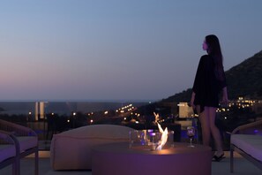 A person standing by a table with a fire feature and a glass of wine, overlooking a cityscape at dusk. - Vitsila Villa II, Design, Sea Views & Family Fun (Rethymno)