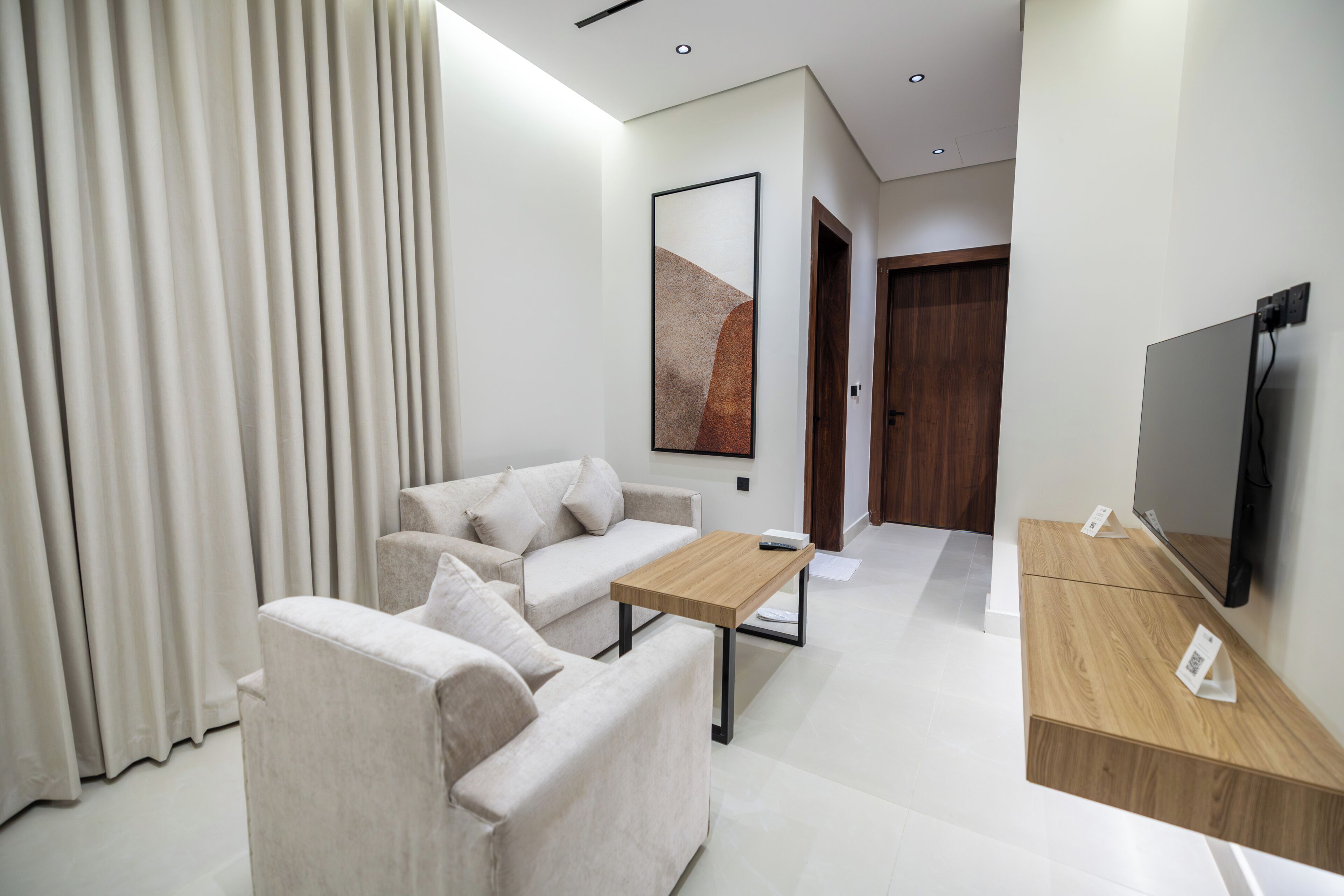 Quarto de hotel moderno com sofá, mesa de cabeceira de madeira, TV de tela plana e uma grande janela com cortinas.