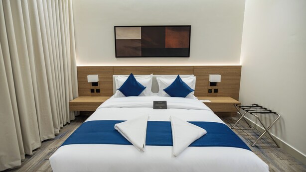 A hotel room with a neatly made bed, a headboard with a light wood finish, two bedside tables with lamps, a wall-mounted artwork, and a window with curtains.