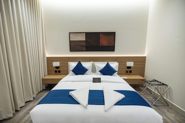 A hotel room with a neatly made bed, a headboard with a light wood finish, two bedside tables with lamps, a wall-mounted artwork, and a window with curtains.