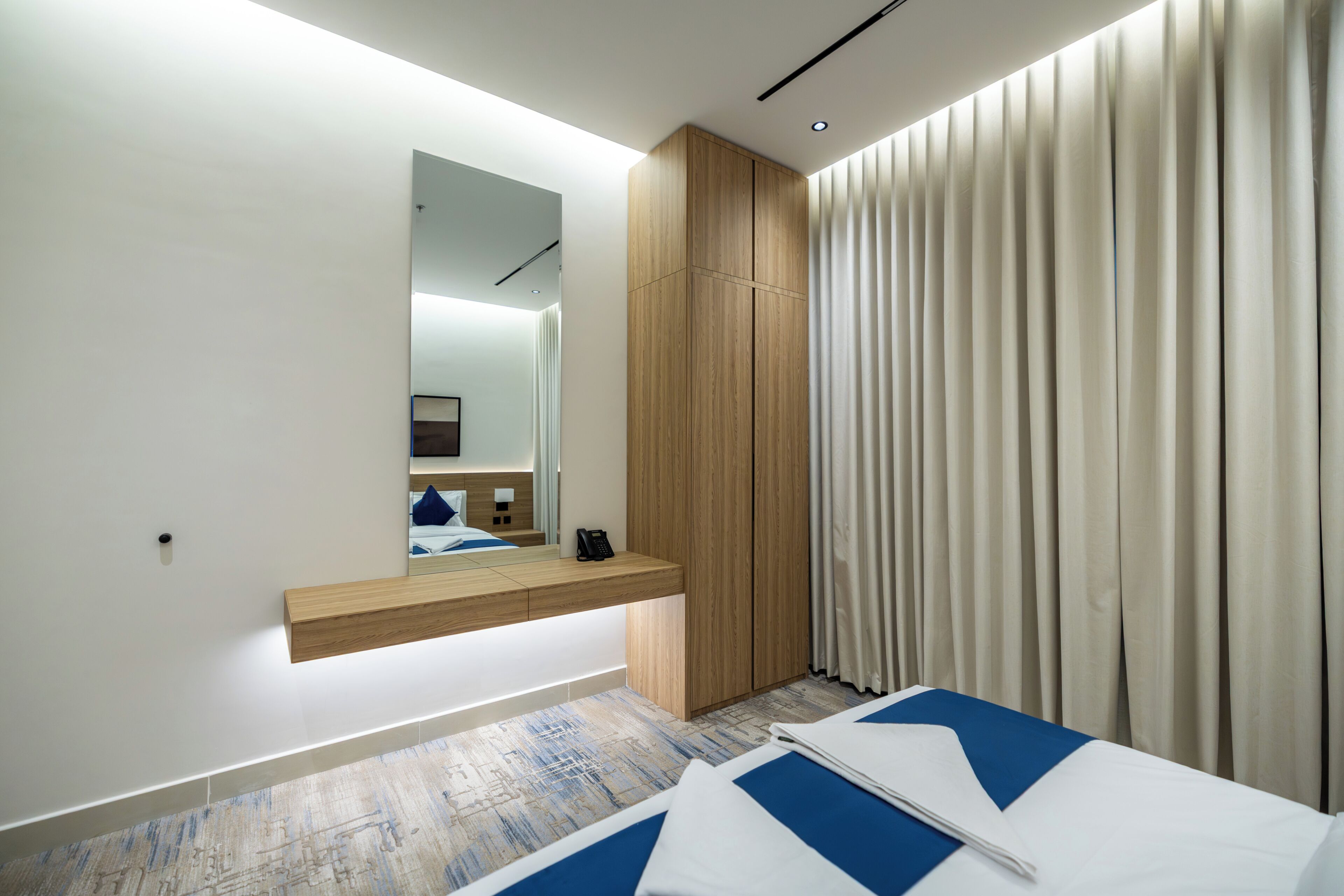 A modern hotel room with a wooden wardrobe, a bedside table with a phone, and a large window with curtains.