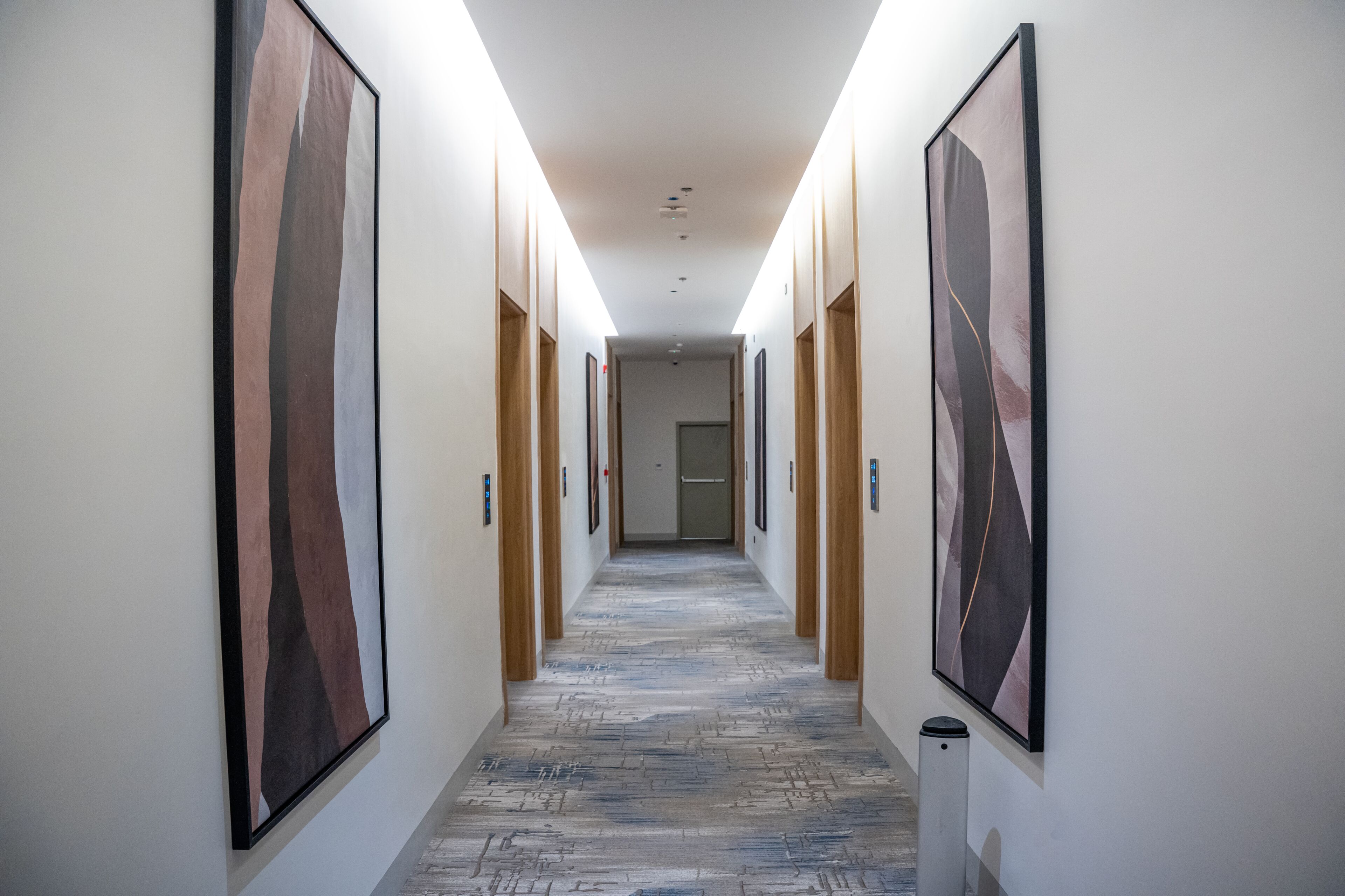 Um corredor com piso estampado, quadros nas paredes e uma lixeira no final.
