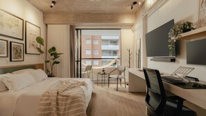 A modern hotel room with a bed, desk, chair, TV, and a view of a building through the window. - Studio Nomad Pro | A/C, just steps from the Metro (Sabaneta)