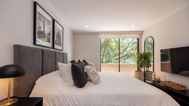 A modern bedroom with a large bed, a bedside table with a lamp, a flat-screen TV, and a window with a view of trees. - Live in Provenza Modern 1 BR Security (Medellín)