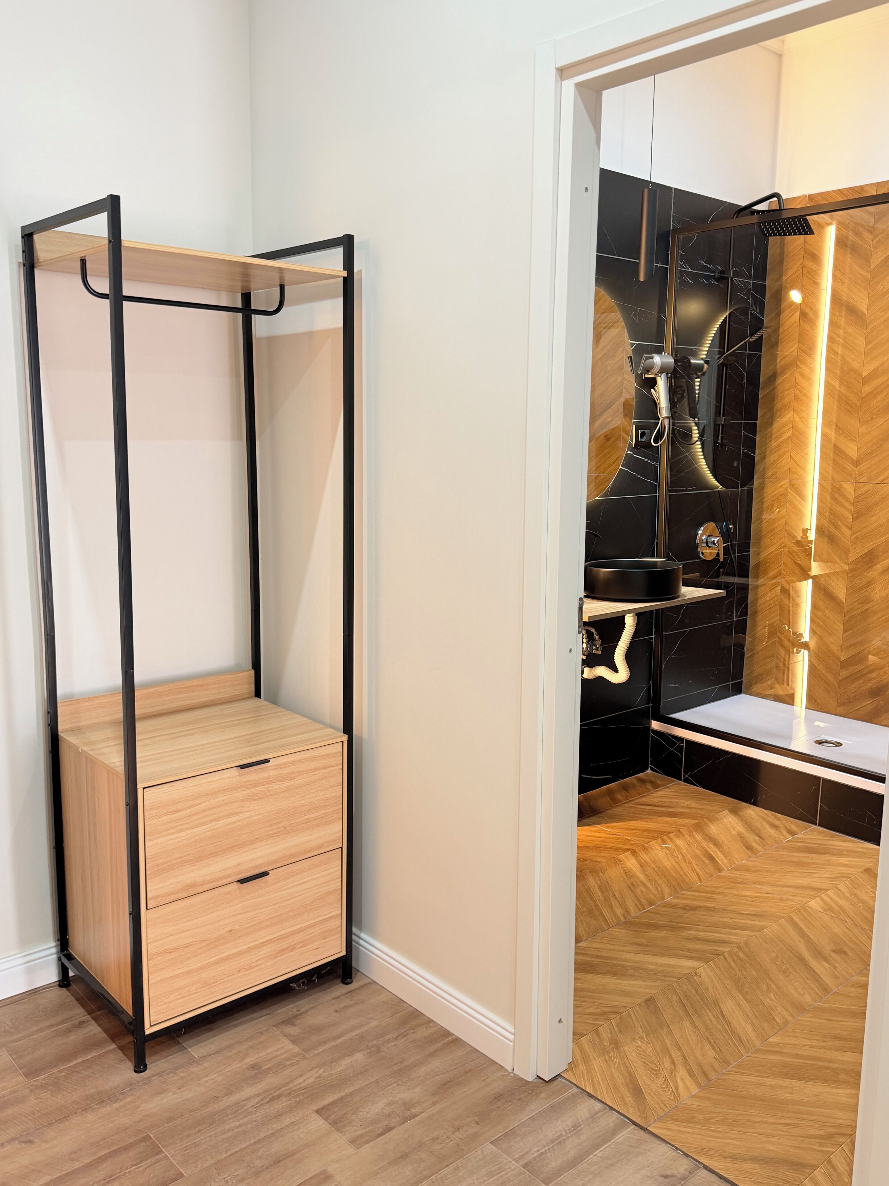 Een kamer met een houten garderobe, een zwart metalen bed en een badkamer met douche en wastafel.