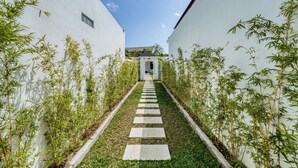 Uma passagem ladeada por paredes brancas e plantas de bambu, que leva a um edifício com telhado escuro.