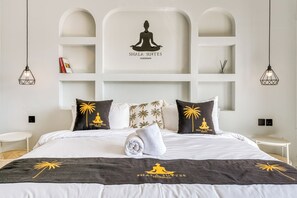 Hotelkamer met een bed, kussens met palmboommotieven, en een wandplank met een mediterende figuur en tekst.