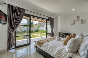 A bedroom with a bed, a desk, and a view of a garden through large windows. - Shala Suites Bali - Pererenan (Mengwi)