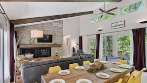 A spacious living area with a fireplace, a dining table set for four, and a large window overlooking greenery. - Whimsical Cabin: Secret Room, Pizza & HOT TUB (Basye)