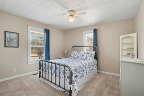 A bedroom with a bed, a ceiling fan, a window with curtains, a bedside table, and a wall-mounted shelf. - Peaceful 4-bedroom cottage in Oyster Bay Golf Community (Sunset Beach)