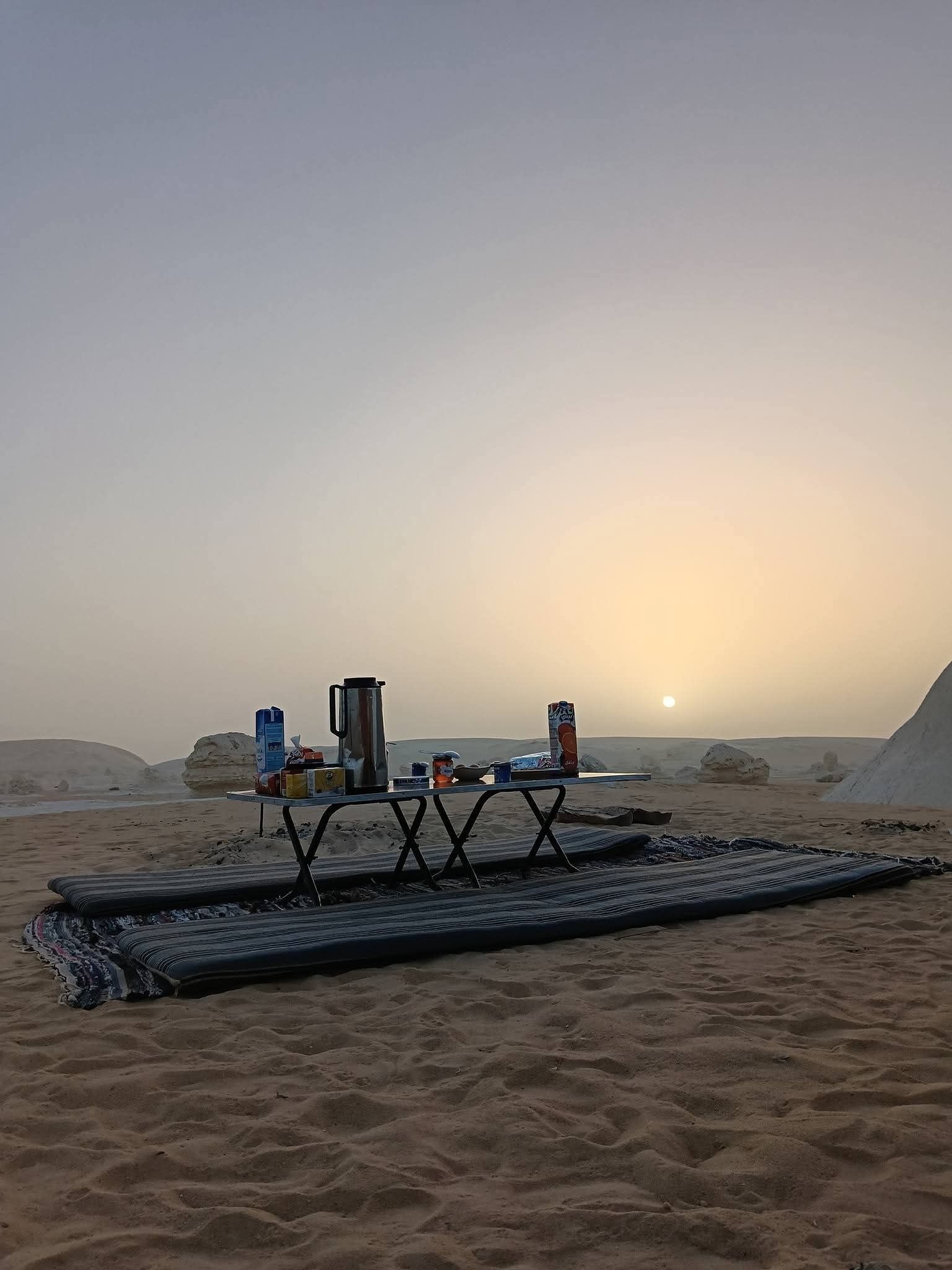 Un campement dans le désert, avec une table dressée pour le repas, entouré de dunes de sable et sous un ciel dégagé.