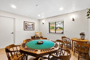 A room with a round wooden table with a green felt top, surrounded by wooden chairs, a window with a view of houses, and a door.