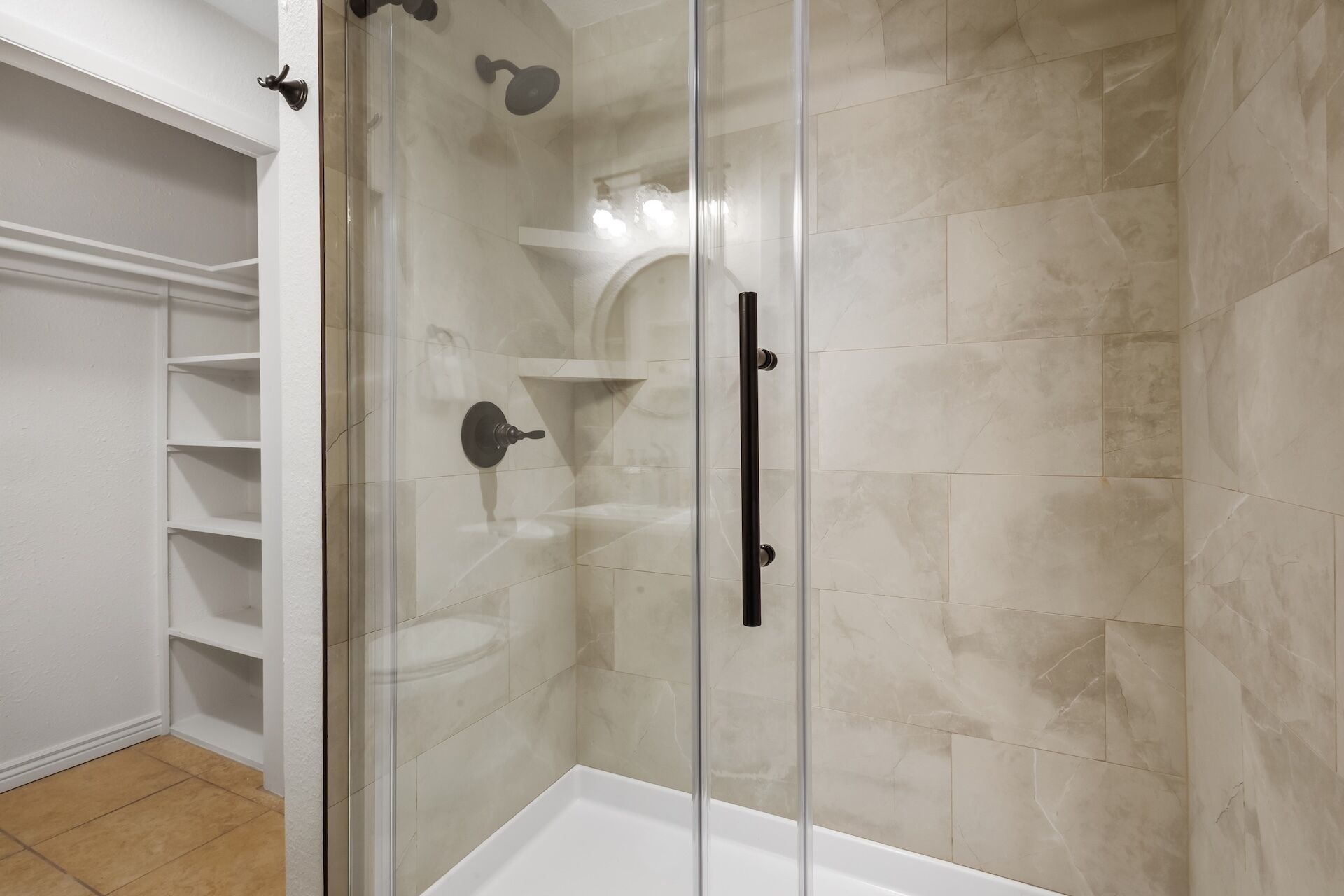 A glass-enclosed shower with a white bathtub, beige marble tiles, and a white shelving unit.
