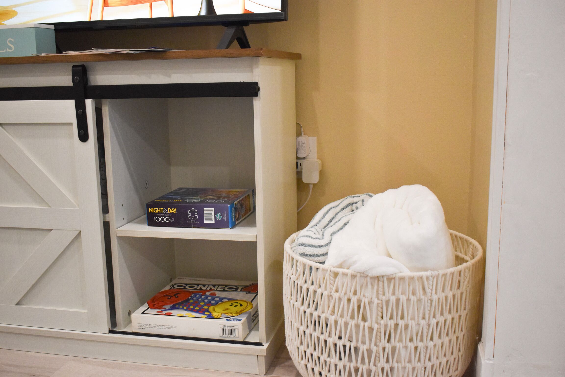 Une pièce avec un meuble de rangement blanc contenant une boîte de jeux de puzzle et un magazine. Un panier en osier blanc est placé contre le mur.