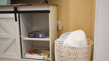 Une pièce avec un meuble de rangement blanc contenant une boîte de jeux de puzzle et un magazine. Un panier en osier blanc est placé contre le mur.