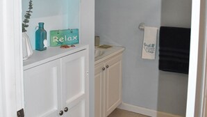 A bathroom with a white door, a shelf with a blue bottle and a 'Relax' sign, a framed picture on the wall, and a towel rack with a black towel.