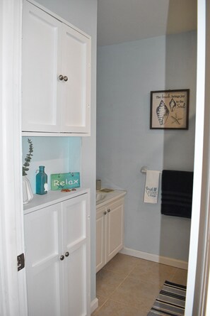 Une salle de bain avec une porte blanche, une étagère sur laquelle se trouve une bouteille bleue et une pancarte indiquant « Détendez-vous », un tableau encadré accroché au mur et un porte-serviettes avec une serviette noire.
