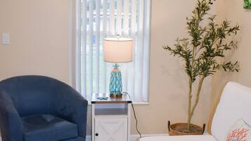 A room with a blue armchair, a white sofa, a lamp on a side table, a potted plant, and a window with blinds.