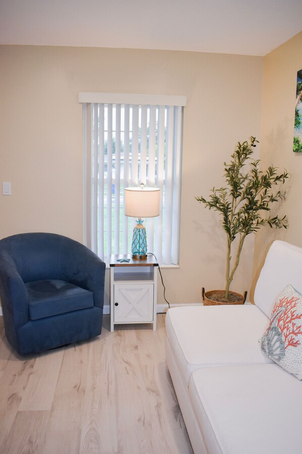 A room with a blue armchair, a white sofa, a lamp on a side table, a potted plant, and a window with blinds.