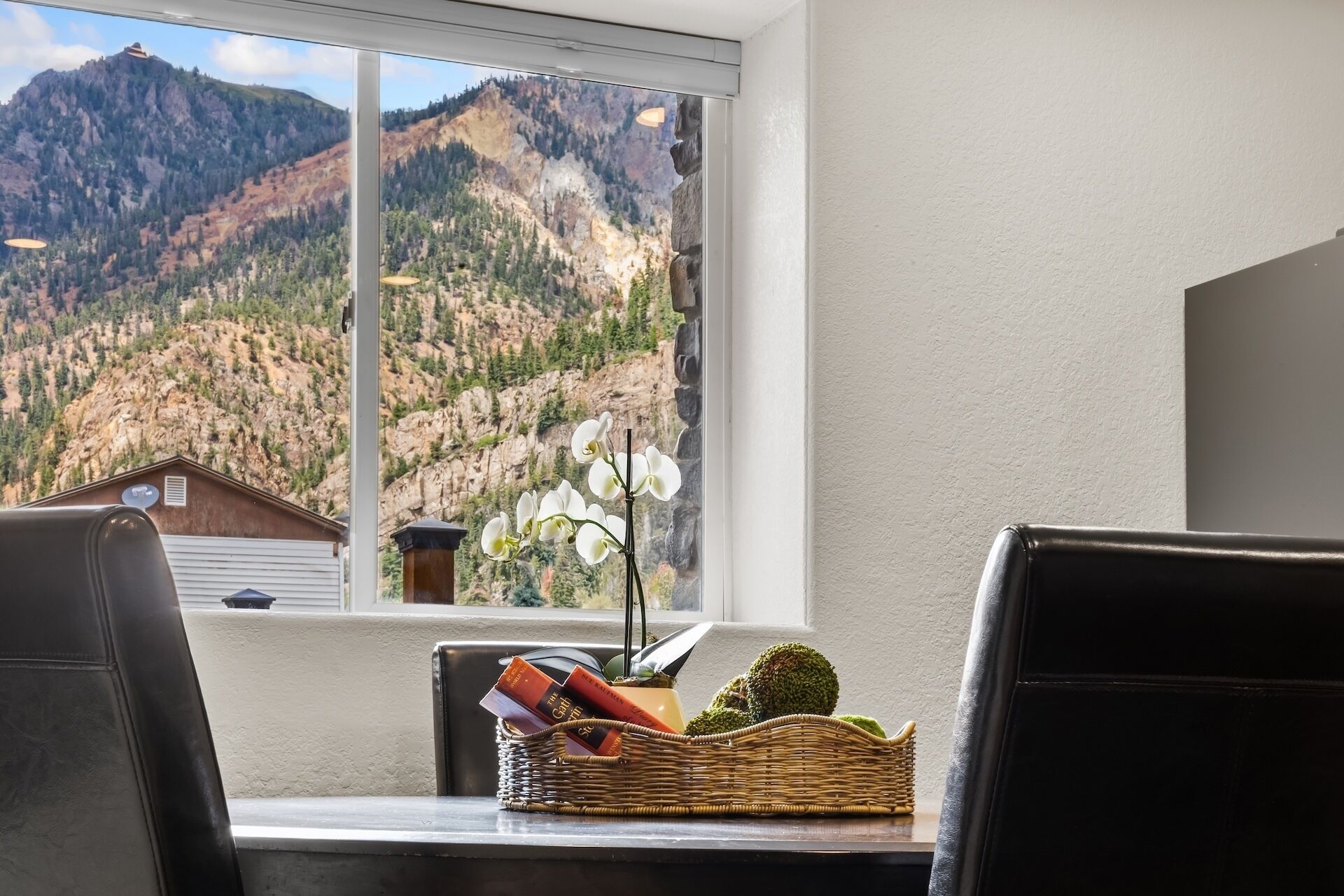 A dining area with a mountain view, a wicker basket with books, and a vase with white flowers.
