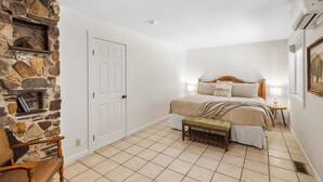 A bedroom with a stone wall, a bed with a wooden headboard, a bench, a bedside table with a lamp, and a framed picture on the wall.