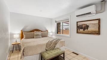 A bedroom with a bed, a bench, a window, an air conditioning unit, and a framed picture on the wall.