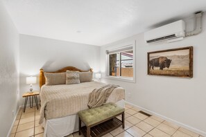Camera da letto con un letto, una panca, una finestra, un condizionatore e un quadro appeso al muro.