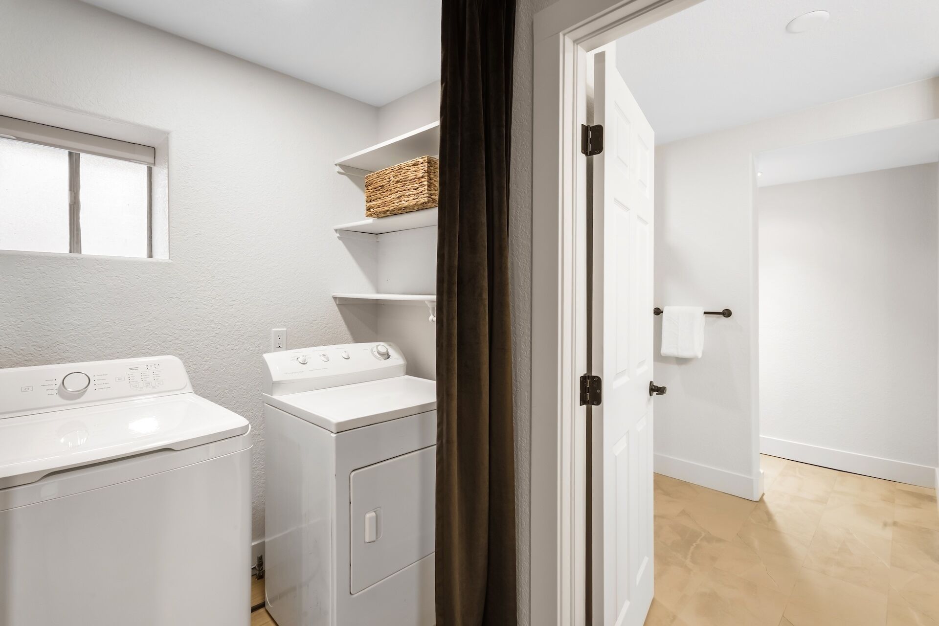 A laundry room with two white washing machines, a shelf with a basket, and a door leading to another room.