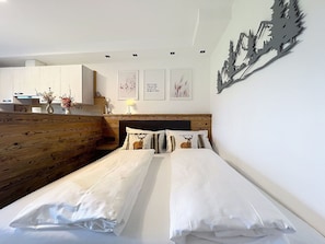 Ein Hotelzimmer mit zwei Betten, einem Holzkopfteil, einer kleinen Küchenzeile und einer Wand mit gerahmten Kunstwerken.
