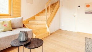 En trætrappe fører op til en etage ovenpå, med en sofa med puder, et lille rundt bord og en væg med et billede.