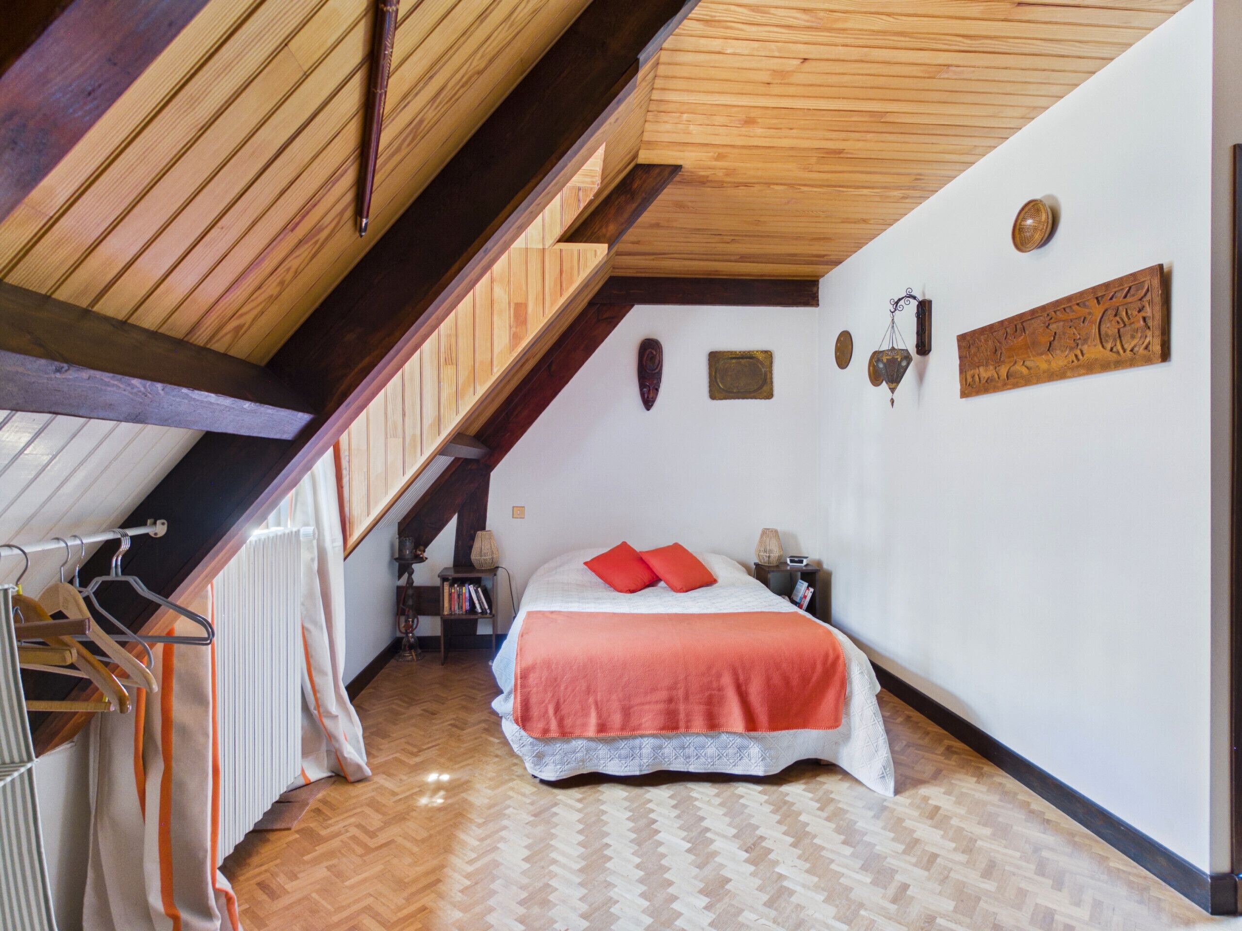 A bedroom with a sloped ceiling, a bed with a red blanket, a wooden headboard, and a small bedside table.