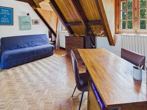 A cozy loft-style room with a herringbone-patterned wooden floor, a blue sofa, a wooden dining table, and a view of greenery through a window. - Fouesnant, family house for 10 with garden (Fouesnant)