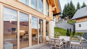 Eine Holzdielen-Terrasse mit Gartenmöbeln, große Schiebeglasfenster, die zu einem Innenraum führen, und Blick auf eine Wohngegend mit Bäumen und einem Gebäude im Hintergrund.